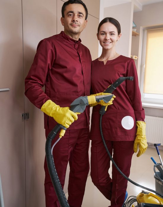 Smiling female cleaner and her calm male colleague with cleaning equipment standing indoors
